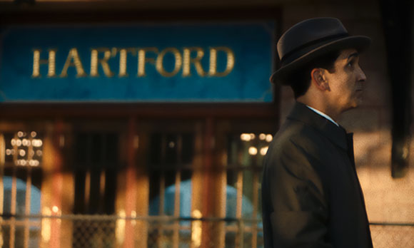 A man in a dark suit and fedora looks to the side in front of a blurred sign that reads HARTFORD.