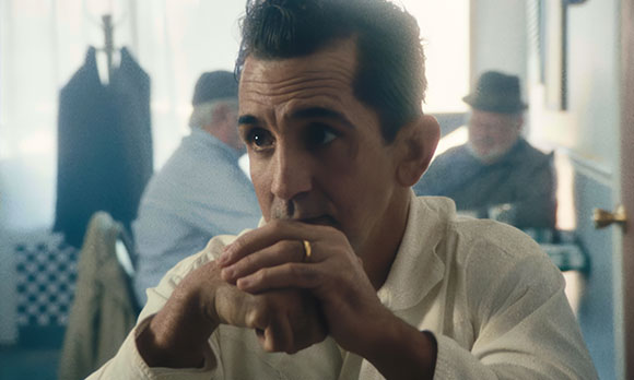A man in a white shirt sits with hands clasped, looking off-camera with a serious expression.