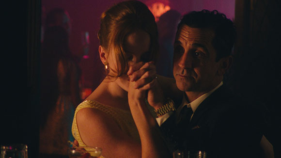 A man and a woman in formal attire hold hands in a dark, moody room with red lighting.