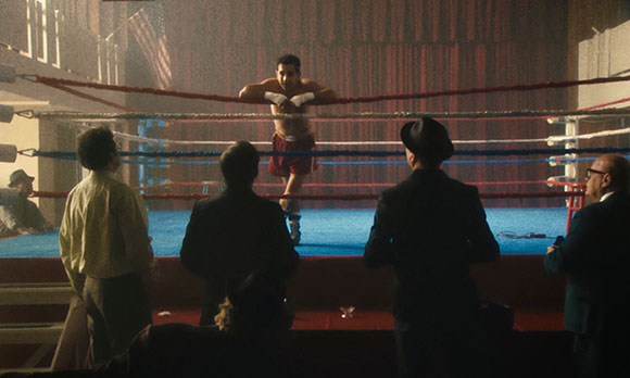 A boxer stands in a blue boxing ring facing four men in suits in a dimly lit arena.