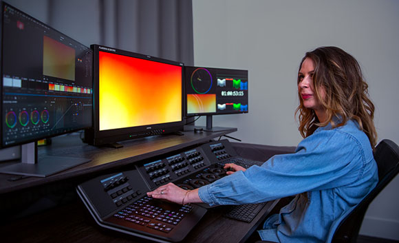 A woman sits at a desk using a control panel and three monitors displaying color grading software.