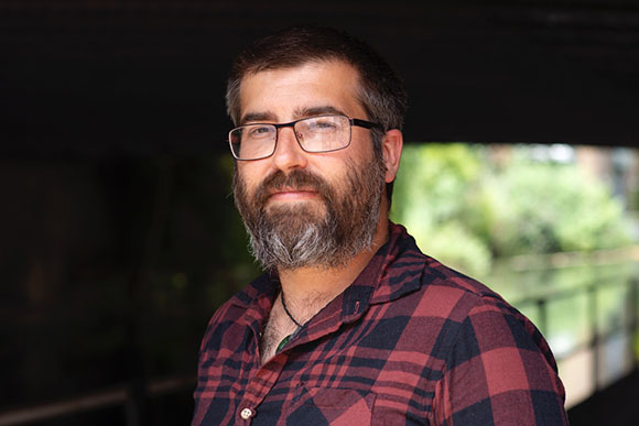 A man with dark hair, a full beard, and glasses wearing a red and black plaid shirt.