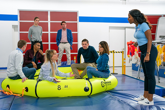 A woman in a blue shirt stands over a group of people sitting in a yellow inflatable life raft.
