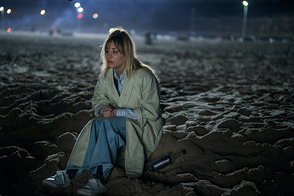A blonde woman in a light trench coat sits on a dark, sandy beach at night under dim lights.