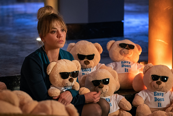 A woman sits looking pensively among several tan teddy bears wearing sunglasses and white t-shirts.
