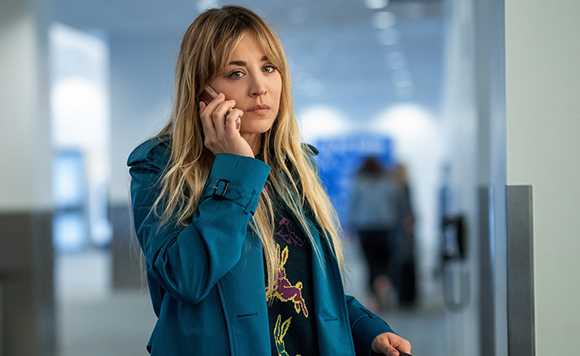 A blonde woman in a teal coat talks on a mobile phone in a bright, blurred hallway.