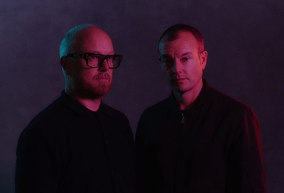 Two men in dark clothing looking forward against a dark purple and blue background.