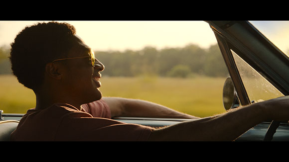 A smiling man wearing sunglasses drives a car through a sunlit field.