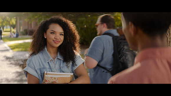 A young woman with voluminous curly hair smiles while looking at a man in a park setting.