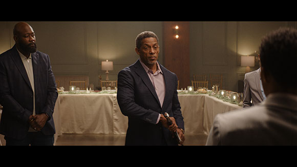 A man in a blue suit holds a handgun while facing another man in a dimly lit dining room.