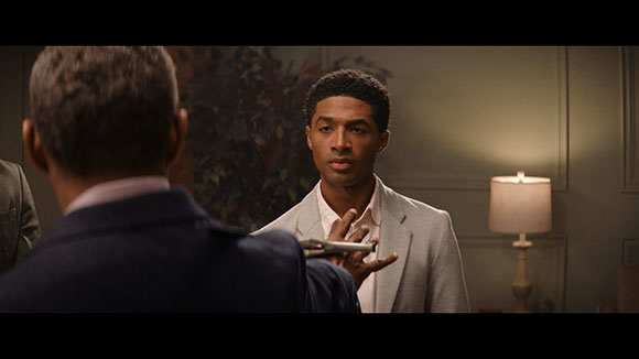 A man in a light grey blazer looks toward a man in a dark suit holding a handgun.