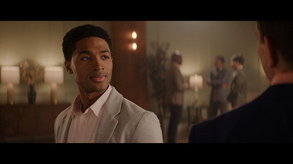 A man in a light grey blazer and pink shirt looks toward a person in profile in a dimly lit room.