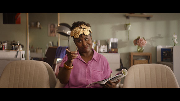 A woman in a pink shirt with gold leaf on her forehead holds an open magazine in a salon.