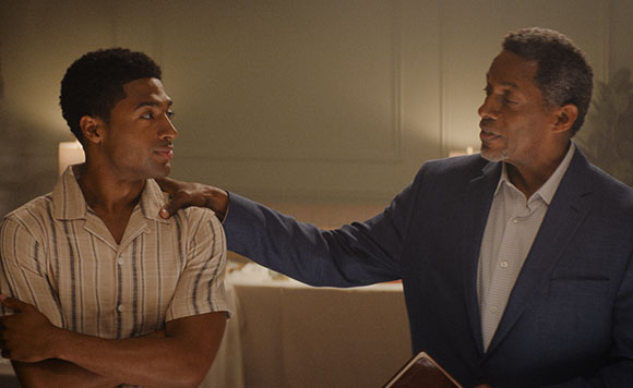 A younger man in a striped shirt looks at an older man in a suit who has a hand on his shoulder.