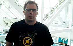 A man with glasses wearing a black t-shirt stands in a bright building with white structural beams.