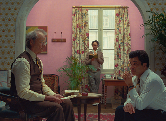 Two men sit in a pink room facing each other while a man stands by a window reading a book.