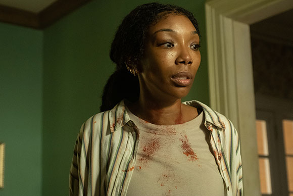 A woman with dark braided hair and blood on her white shirt and neck looks on with a startled expression.