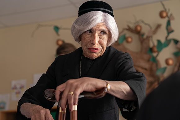 An elderly woman with short grey hair wearing a black garment looks thoughtfully to the side.
