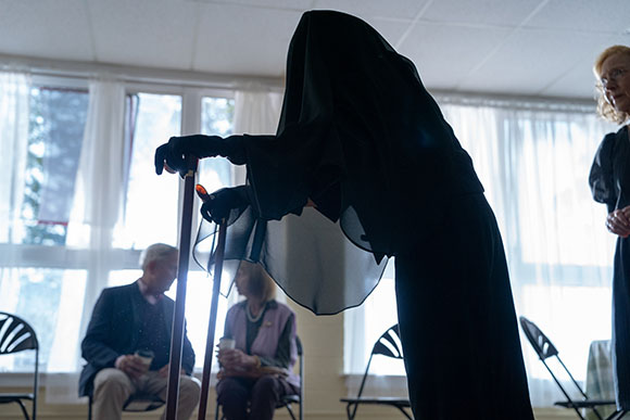 A person in a black hooded cloak leans forward using two walking canes in a bright room.