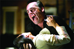 A man in a black shirt screams with an open mouth while leaning over a person in a light-colored jacket.