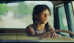A young girl with dark braided hair looks out of a car window.