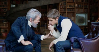 An older man with a grey beard leans toward a younger man sitting in a leather chair in a library.
