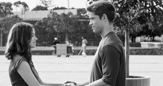 A young man and woman stand facing each other, holding hands in an outdoor plaza.