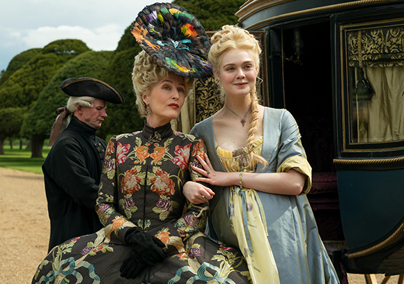 Two women in elaborate 18th-century gowns stand next to a dark carriage in a garden setting.
