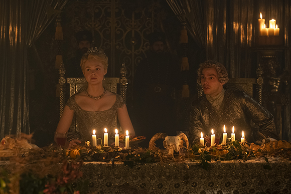 A woman in a crown and a man in period clothing sit at a long table lit by many candles.
