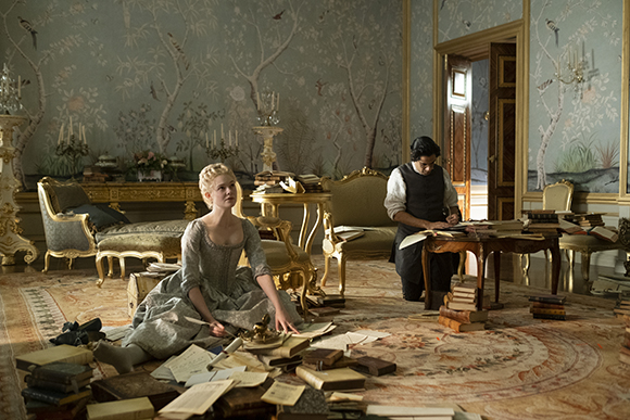 A woman in a grey period dress sits on a patterned rug surrounded by open books in a grand room.