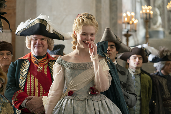 A blonde woman in a light blue period gown smiles while men in 18th-century military and formal attire stand behind her.