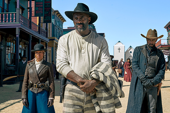 Three people in period clothing stand on a dirt street in a Western town with colorful buildings.