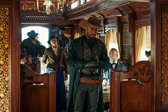 People in 19th-century attire stand and sit inside a richly carved wooden train car.
