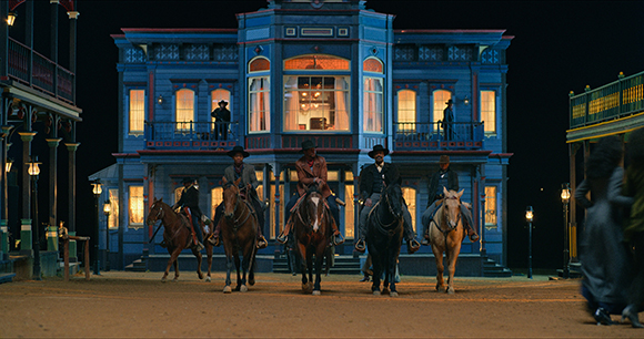 Five people riding horses across a dirt street in front of a large, two-story blue building at night.