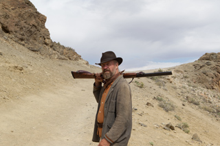 A bearded man in a brown hat and grey jacket carries a long rifle over his shoulder in a rocky desert.