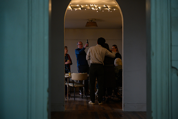 A group of people stand around a table in a dimly lit room viewed through an open doorway.