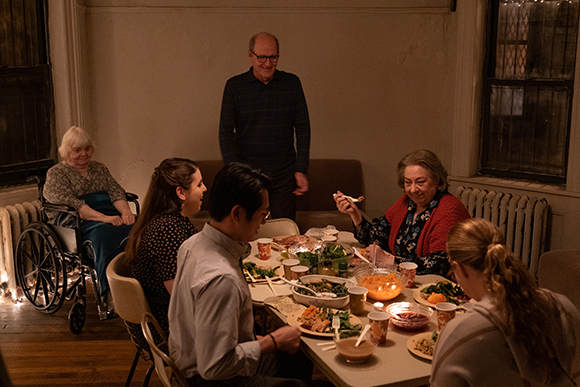 A group of people sit around a dining table filled with food in a dimly lit room.