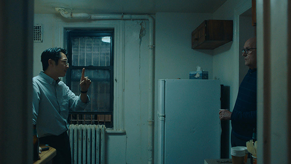 A man in a light shirt gestures with one finger raised while talking to an older man near a refrigerator.