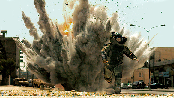 A person in heavy protective gear runs toward the camera as a large explosion erupts behind them.