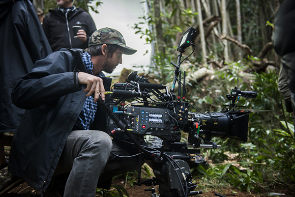 A man in a camouflage cap and dark jacket adjusts a large professional cinema camera in a forest.