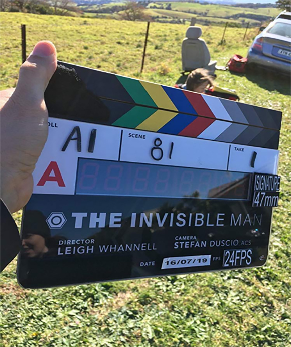 A hand holds a film clapperboard for 'The Invisible Man' against a background of a grassy field and hills.