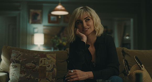 A blonde woman in a black shirt sits on a couch, smiling with her hand on her chin in a dimly lit room.