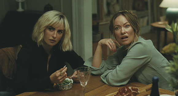 Two women sit at a wooden table; one with blonde hair looks forward, the other leans in with a surprised expression.
