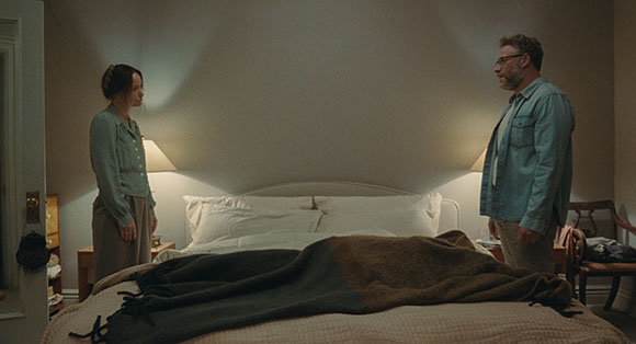A man and a woman stand facing each other on opposite sides of a large bed in a dimly lit room.