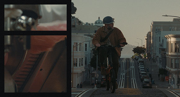 A man in a helmet and brown jacket rides a bicycle down a steep urban hill toward the camera.