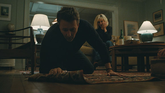 A man crawls across a patterned rug in a dark living room as a woman sits on a sofa in the background.