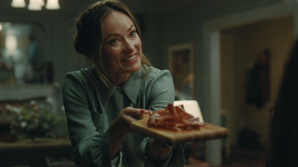 A smiling woman in a green blouse holds out a wooden platter with thin slices of meat.