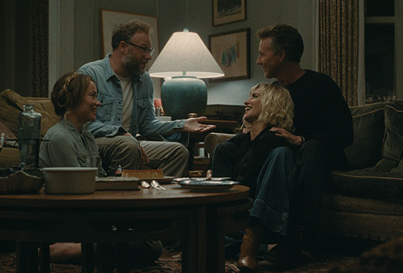Four people sit together in a dimly lit living room around a coffee table with a lamp.