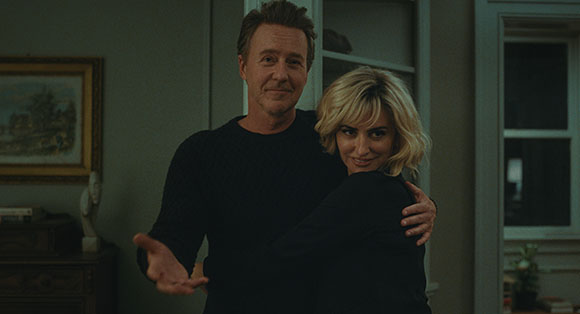 A man in a black sweater smiles while embracing a blonde woman in a dark room.