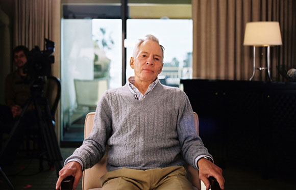 A man in a grey sweater sits in a chair while a camera operator films him in a room.